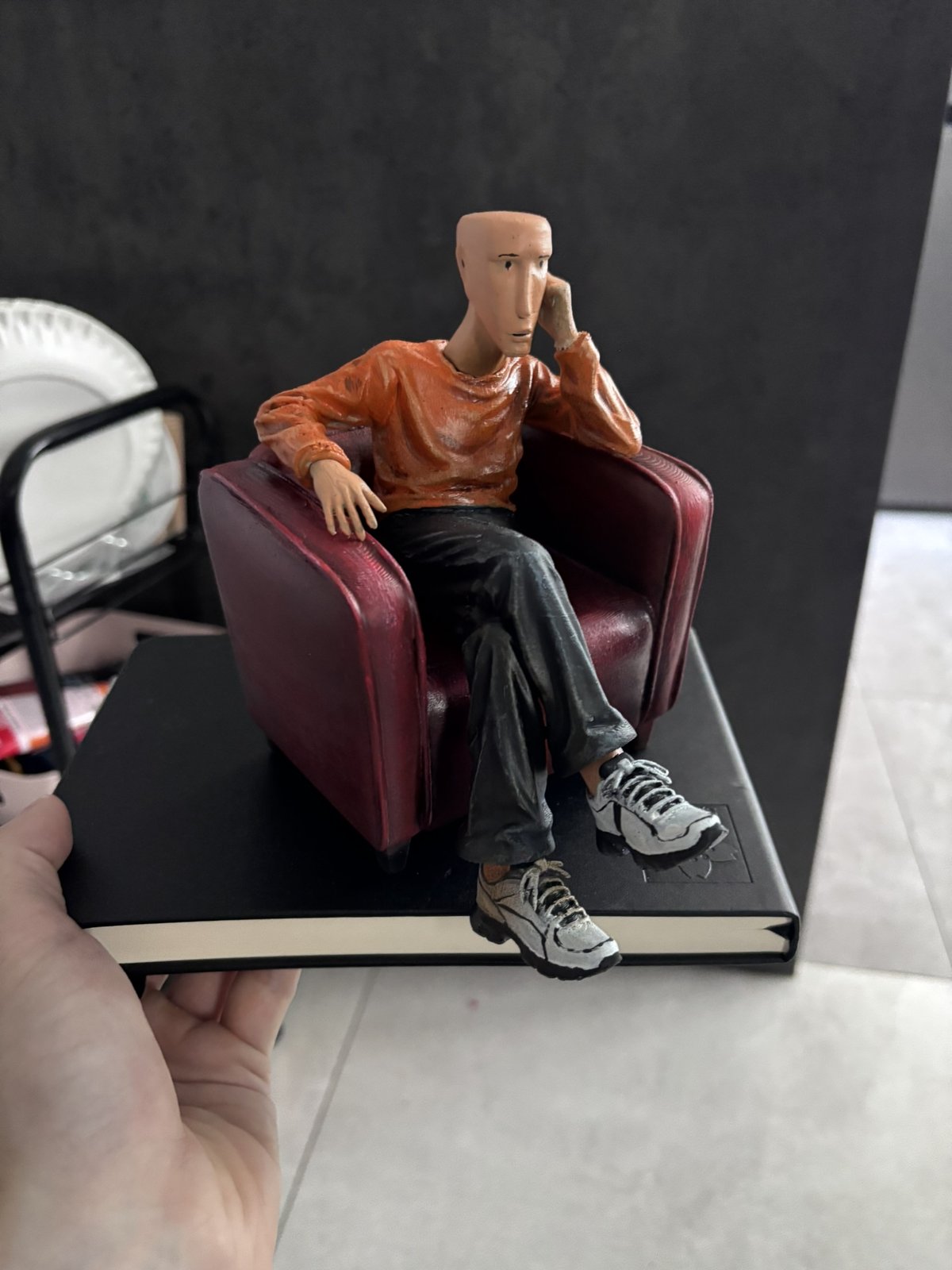 The Host sculpture: a painted 3D-printed figure of a man in an orange sweater sitting in a burgundy armchair